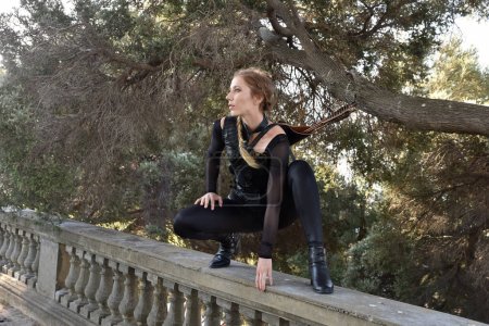 Portrait of beautiful female model with blonde plait, wearing black leather catsuit costume, fantasy assassin warrior.  Crouching sitting pose on stone  balcony of  castle background 