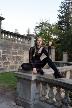 Portrait de belle femme modèle avec tresse blonde, portant costume de catsuit en cuir noir, guerrier assassin fantaisie. Pose assise accroupie sur le balcon en pierre de fond du château 