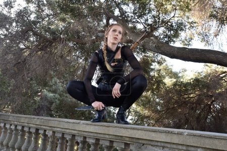 Portrait of beautiful female model with blonde plait, wearing black leather catsuit costume, fantasy assassin warrior.  Crouching sitting pose, holding knife on stone  balcony of  castle background