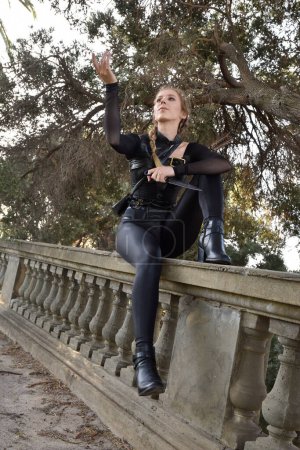Portrait of beautiful female model with blonde plait, wearing black leather catsuit costume, fantasy assassin warrior.  Crouching sitting pose, holding knife on stone  balcony of  castle background