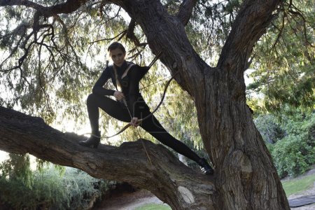 Foto de Full length portrait of beautiful female model with blonde plait, wearing black leather catsuit costume, fantasy assassin warrior. holding bow and arrow. Posing in forest  tree branch - Imagen libre de derechos