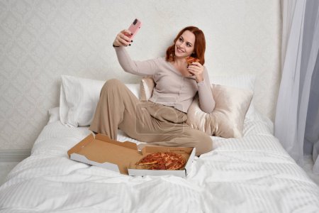 Photo for Portrait of beautiful red haired woman wearing comfortable pyjamas.Relaxing at home, eating  take out pizza,  in a  glamorous bedroom background. - Royalty Free Image