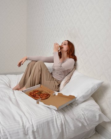 Photo for Portrait of beautiful red haired woman wearing comfortable pyjamas.Relaxing at home, eating  take out pizza,  in a  glamorous bedroom background. - Royalty Free Image