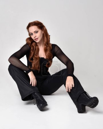 Full length portrait of beautiful woman with long red hair wearing sheer corset top and leather pants.  Sitting pose with gestural hands reaching out, kneeling on floor. Isolated on white studio background.