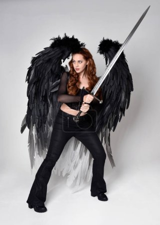 Full length portrait of beautiful woman with long red hair wearing  corset top, leather pants, large black angel feather wings. Standing pose holding sword weapon, walking forwards with gestural hands reaching out. Isolated on white studio background