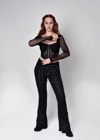 Full length portrait of beautiful woman with long red hair wearing black corset top and leather pants. Standing pose, walking forwards with gestural hands reaching out. Isolated on white studio background.