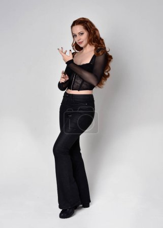 Full length portrait of beautiful woman with long red hair wearing black corset top and leather pants. Standing pose, walking forwards with gestural hands reaching out. Isolated on white studio background.
