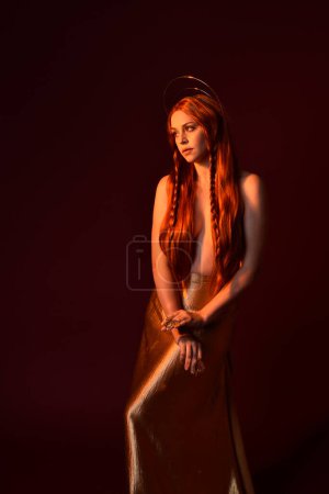 Nahaufnahme Fantasie-Porträt der schönen Frau Modell mit roten Haaren, Göttin Seidengewänder & goldene Krone. Posieren mit ausgestreckten Händen, isoliert auf dunkelrotem Studiohintergrund 