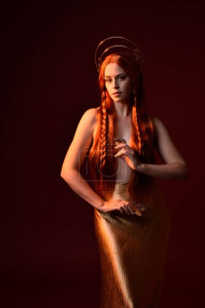 Photo for Close up fantasy portrait of beautiful woman model with red hair, goddess silk robes & gold crown.  Posing with gestural hands reaching out, isolated on dark red studio background - Royalty Free Image