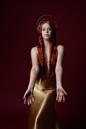 Photo for Close up fantasy portrait of beautiful woman model with red hair, goddess silk robes & gold crown.  Posing with gestural hands reaching out, isolated on dark red studio background - Royalty Free Image