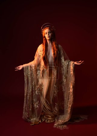 Photo for Full length fantasy portrait of beautiful woman model with red hair, goddess silk robes & gold crown. Standing pose gestural hands reaching out isolated on dark red studio background - Royalty Free Image