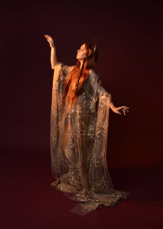 Portrait fantaisie pleine longueur de belle femme modèle aux cheveux roux, robes de soie déesse & couronne d'or. Pose debout mains gestuelles tendues isolées sur fond de studio rouge foncé 