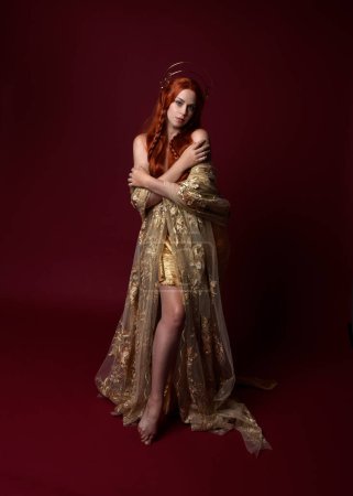 Photo for Full length fantasy portrait of beautiful woman model with red hair, goddess silk robes & gold crown. Standing pose gestural hands reaching out isolated on dark red studio background - Royalty Free Image