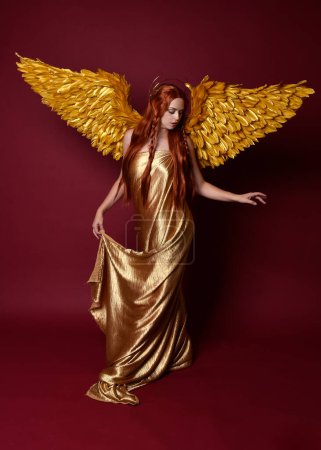 Portrait complet de la belle femme modèle avec de longs cheveux rouges, robes de soie or, couronne & ailes fantaisie ange plume. Pose debout mains gestuelles tendues isolées sur fond de studio rouge foncé