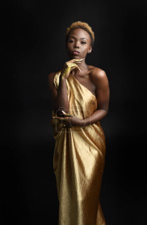  Fantasie-Porträt der schönen afrikanischen Frau Modell mit Afro, Göttin Seide Roben und reich verzierten Blumenkranz Krone. Gestenreich posiert er mit goldenen Blumen. isoliert auf dunklem Studiohintergrund 