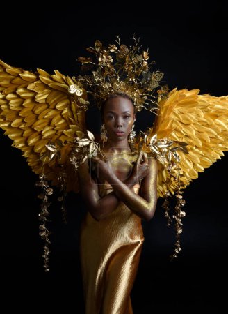 Photo for Fantasy portrait of beautiful african woman model with afro, goddess silk robes, ornate crown & gold angel wings.  gestural Posing holding golden flowers., isolated on dark  studio background - Royalty Free Image