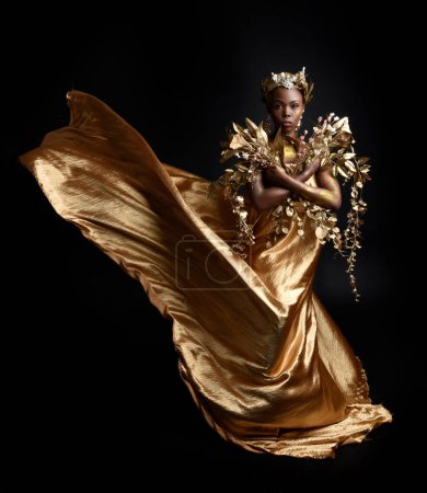  Fantasie-Porträt der schönen afrikanischen Frau Modell mit Afro, Göttin Seide Roben und reich verzierten Blumenkranz Krone. Gestenreich posiert er mit goldenen Blumen. isoliert auf dunklem Studiohintergrund 