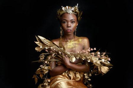  portrait fantaisiste de belle femme africaine modèle avec afro, robes de soie de déesse et couronne de couronne de couronne fleurie ornée. pose gestuelle tenant des fleurs dorées. isolé sur fond sombre studio 
