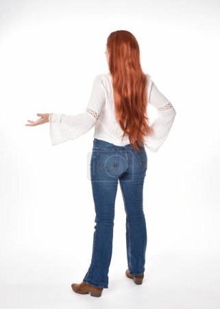 Foto de Retrato de longitud completa de modelo de mujer hermosa con el pelo largo y rojo, el uso de ropa casual blusa blanca superior y jeans de mezclilla, aislado en fondo de estudio blanco. Posar hacia atrás, alejarse de la cámara. - Imagen libre de derechos