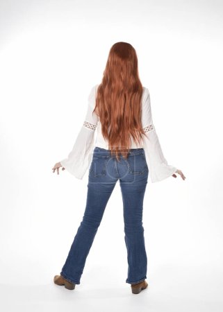 Foto de Retrato de longitud completa de modelo de mujer hermosa con el pelo largo y rojo, el uso de ropa casual blusa blanca superior y jeans de mezclilla, aislado en fondo de estudio blanco. Posar hacia atrás, alejarse de la cámara. - Imagen libre de derechos
