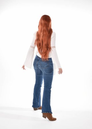 Ganzkörperporträt der schönen Frau Modell mit langen roten Haaren, trägt legeres Outfit weiße Bluse Top und Jeans, isoliert auf weißem Studiohintergrund. Rückwärts stehend posieren, von der Kamera weggehen.