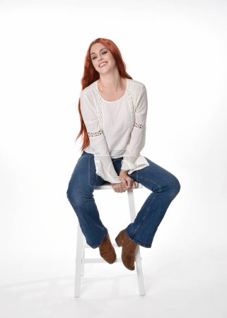 Foto de Retrato de longitud completa de modelo de mujer hermosa con el pelo largo y rojo, el uso de ropa casual blusa blanca superior y jeans de mezclilla, aislado en fondo de estudio blanco. Postura sentada relajada, sentada en silla con poses de mano gestual. - Imagen libre de derechos