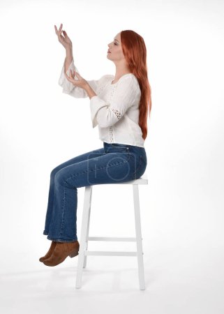 Foto de Retrato de longitud completa de modelo de mujer hermosa con el pelo largo y rojo, el uso de ropa casual blusa blanca superior y jeans de mezclilla, aislado en fondo de estudio blanco. Postura sentada relajada, sentada en silla con poses de mano gestual. - Imagen libre de derechos