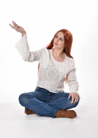Foto de Retrato de longitud completa de modelo de mujer hermosa con el pelo largo y rojo, el uso de ropa casual blusa blanca superior y jeans de mezclilla, aislado en fondo de estudio blanco. Postura sentada relajada, sentada en el suelo con posturas de mano gestual. - Imagen libre de derechos