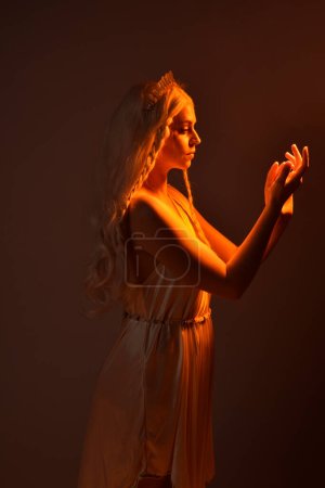 Photo for Portrait of beautiful blonde woman wearing a fantasy goddess toga costume with  crystal crown.Backlit a glowing isolated studio background with  orange silhouette lighting. - Royalty Free Image
