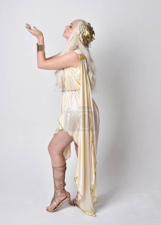 Photo for Full length portrait of beautiful blonde woman wearing a fantasy goddess toga costume with  magical crown.Standing pose, facing backwards away from camera.  isolated on white studio background. - Royalty Free Image