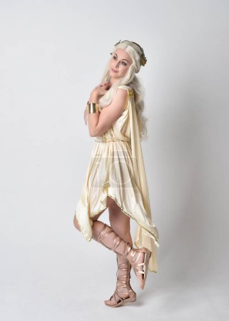 Full length portrait of beautiful blonde woman wearing a fantasy goddess toga costume with  magical crown.Standing, dancing pose with gestural hands reaching out, isolated on white studio background.