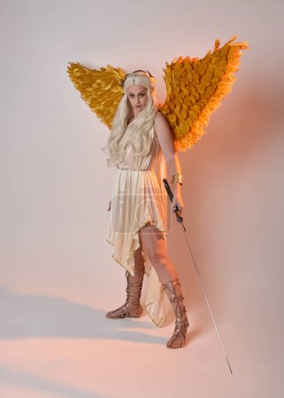 Full length portrait of beautiful blonde woman wearing a fantasy goddess toga costume with feathered angel wings, holding a sword weapon. Jumping pose like flying, isolated on white studio background.