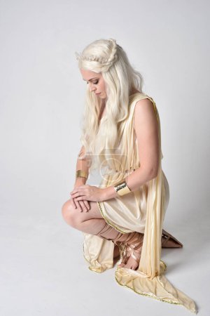 Photo for Full length portrait of beautiful blonde woman wearing a fantasy goddess toga costume with  magical crown.Seated pose, sitting on floor. isolated on white studio background. - Royalty Free Image
