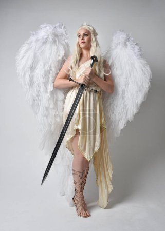 Ganztägiges Porträt der schönen blonden Frau in einem Fantasie-Toga-Kostüm mit gefiederten Engelsflügeln und einer Schwertwaffe. Springende Pose wie im Flug, isoliert auf weißem Studiohintergrund.