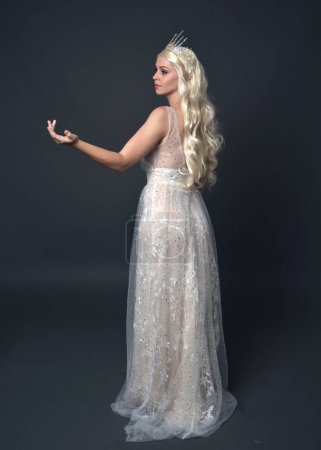 Photo for Full length portrait of beautiful women with long blonde hair, wearing fantasy  princess crown and elegant white ball gown, standing pose with hand gesture. Isolated on dark grey studio background. - Royalty Free Image