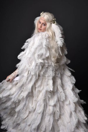 Photo for Fantasy portrait of beautiful female model with long blond hair wearing otherworldly  white feathered cloak costume and headdress, isolated on dark studio background. - Royalty Free Image