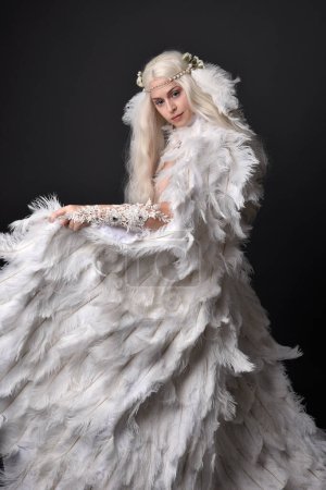 Fantasie-Porträt der schönen weiblichen Modell mit langen blonden Haaren trägt jenseitige weiß gefiederten Mantel Kostüm und Kopfbedeckung, isoliert auf dunklem Studiohintergrund.