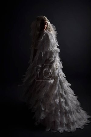 Foto de Retrato de fantasía de hermosa modelo femenina con el pelo largo y rubio con traje de capa de plumas blanco de otro mundo y tocado, aislado en el fondo oscuro del estudio. - Imagen libre de derechos