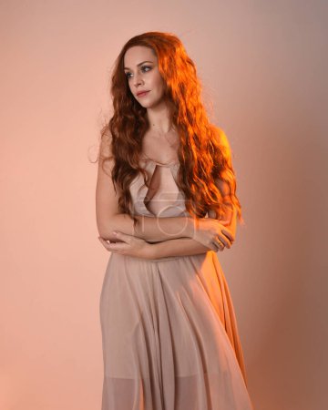 Close up portrait of beautiful brunette  female model wearing a cream dress with golden back light lighting.Gestural arm poses with hands reaching out, isolated on studio background.