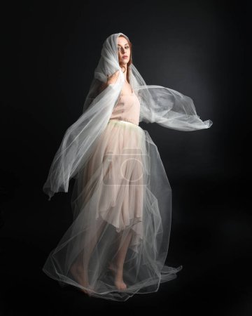 Foto de Retrato de longitud completa de la hermosa mujer con vestido blanco vestido con tela velada fantasmal que fluye, aislado en el fondo oscuro del estudio. - Imagen libre de derechos