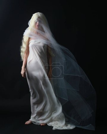 Foto de Retrato de longitud completa de la hermosa mujer rubia con vestido blanco vestido con tela velada fantasmal que fluye, aislado en el fondo del estudio dar. - Imagen libre de derechos
