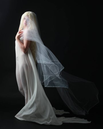 Foto de Retrato de longitud completa de la hermosa mujer rubia con vestido blanco vestido con tela velada fantasmal que fluye, aislado en el fondo del estudio dar. - Imagen libre de derechos