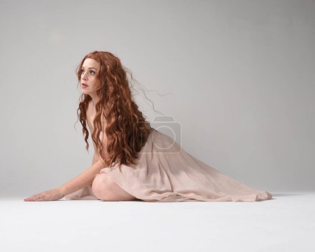 Portrait pleine longueur d'une belle mannequin brune portant une robe rose. pose assise gracieuse, agenouillée sur le sol mains gestuelles. prise de vue en angle bas, isolée sur fond de studio blanc.
