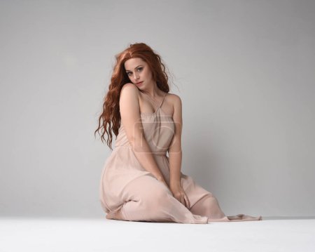 Full length portrait of beautiful brunette model  wearing a  pink dress. graceful sitting  pose, kneeling on floor gestural hands. shot from low angle perspective,  isolated on white studio background.