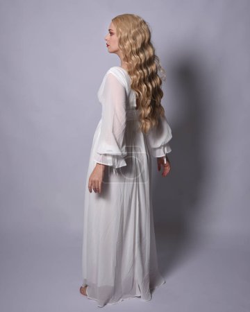 Ganztägiges Porträt einer blonden Frau im weißen historischen Brautkleid. Stehende Pose, nach hinten gewandt, sich von der Kamera entfernend, isoliert auf Studiohintergrund.