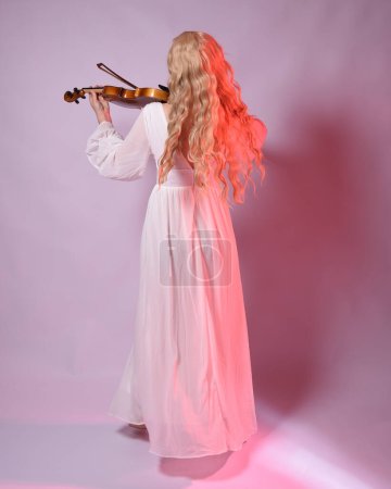 Portrait pleine longueur d'un beau modèle blond portant une élégante robe blanche d'Halloween, personnage de fantaisie historique. face vers l'arrière tenant un instrument de musique pour violon, isolé sur fond de studio.