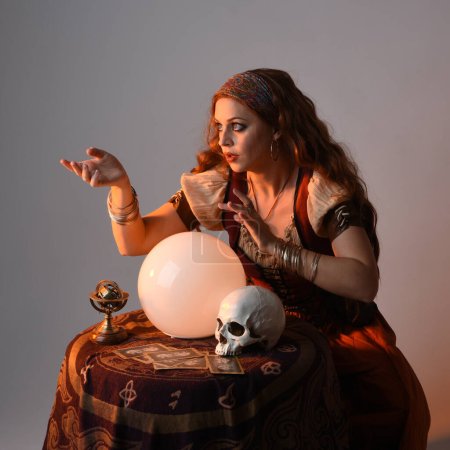 close up portrait of beautiful red haired woman wearing a medieval fantasy fortune teller costume, looking into crystal ball reading the future at seance table. isolated on studio background with cinematic lighting.