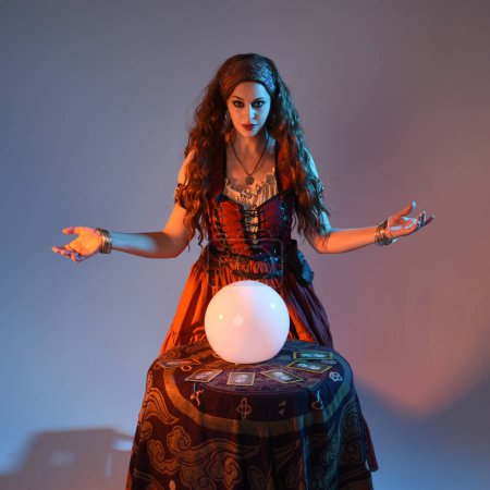 close up portrait of beautiful red haired woman wearing a medieval fantasy fortune teller costume, looking into crystal ball reading the future at seance table. isolated on studio background with moody cinematic lighting.