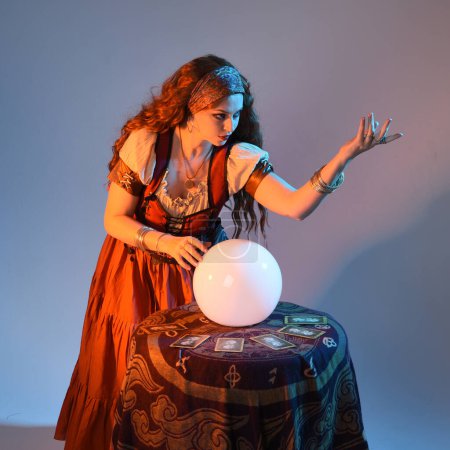 Photo for Close up portrait of beautiful red haired woman wearing a medieval fantasy fortune teller costume, looking into crystal ball reading the future at seance table. isolated on studio background with moody cinematic lighting. - Royalty Free Image