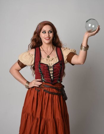 close up portrait of beautiful red haired woman wearing a medieval maiden, fortune teller costume. Posing while  holding a  crystal ball. isolated on studio background.